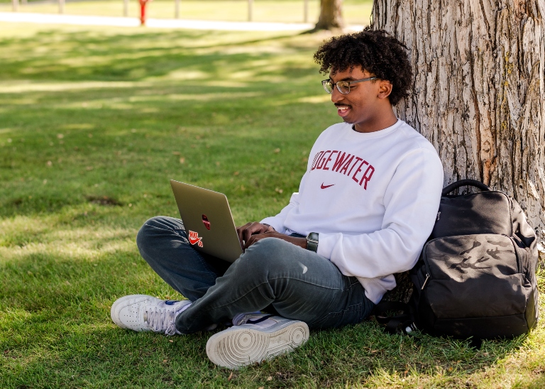 Male student sitting on lawn against a tree while using his laptop.